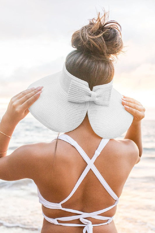Woman at the beach facing the ocean, holding her Foldable Wide Brim Bow Visor, with a crossed back swimsuit.