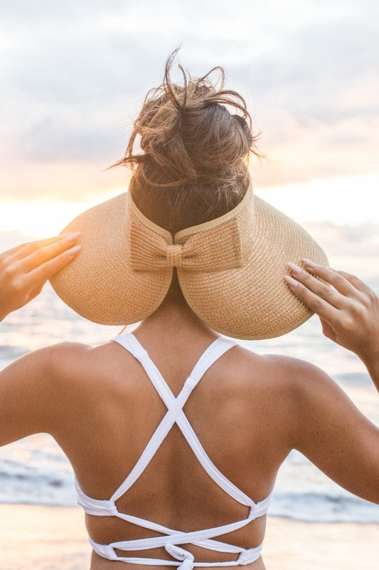 Woman at the beach facing the ocean, holding her Foldable Wide Brim Bow Visor, with a crossed back swimsuit.
