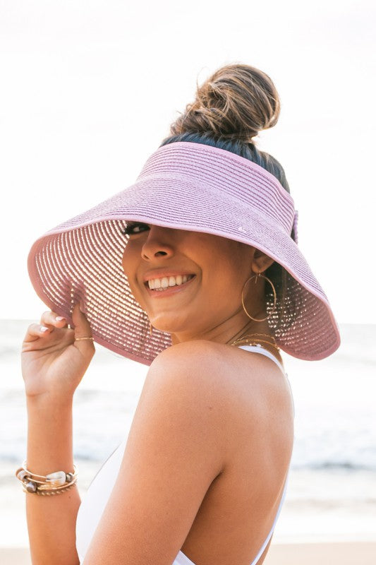 Woman at the beach facing the ocean, holding her Foldable Wide Brim Bow Visor, with a crossed back swimsuit.