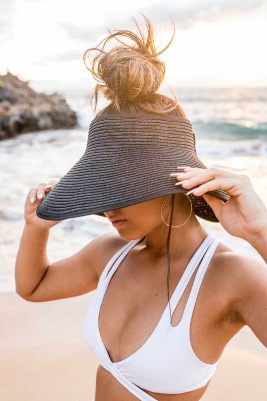 Woman at the beach facing the ocean, holding her Foldable Wide Brim Bow Visor, with a crossed back swimsuit.