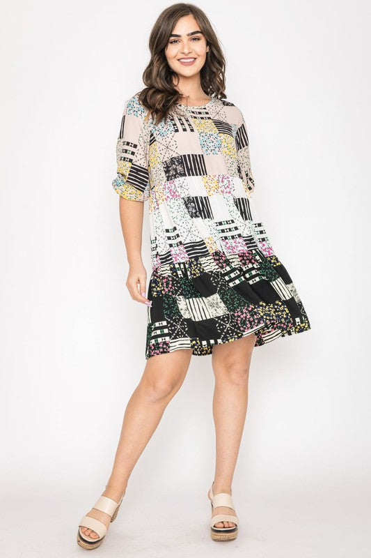 A woman in a Ditsy Floral Patch Work Dress, made in the United States, stands in a cozy living room adorned with lush potted plants and a round wooden table in the background.