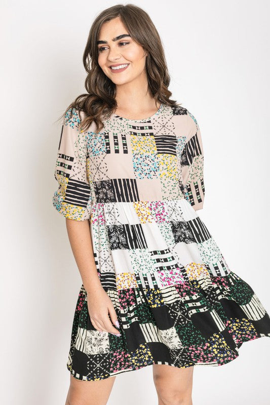 A woman in a Ditsy Floral Patch Work Dress, made in the United States, stands in a cozy living room adorned with lush potted plants and a round wooden table in the background.