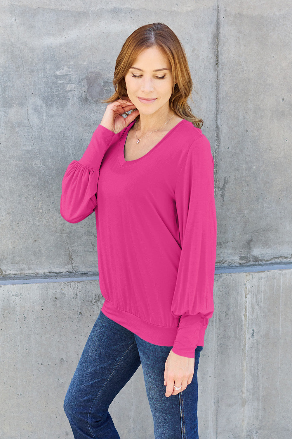 A woman with brown hair is standing against a concrete wall, wearing the Basic Bae Full Size V-Neck Lantern Sleeve Top in teal and blue basic-style jeans, smiling.