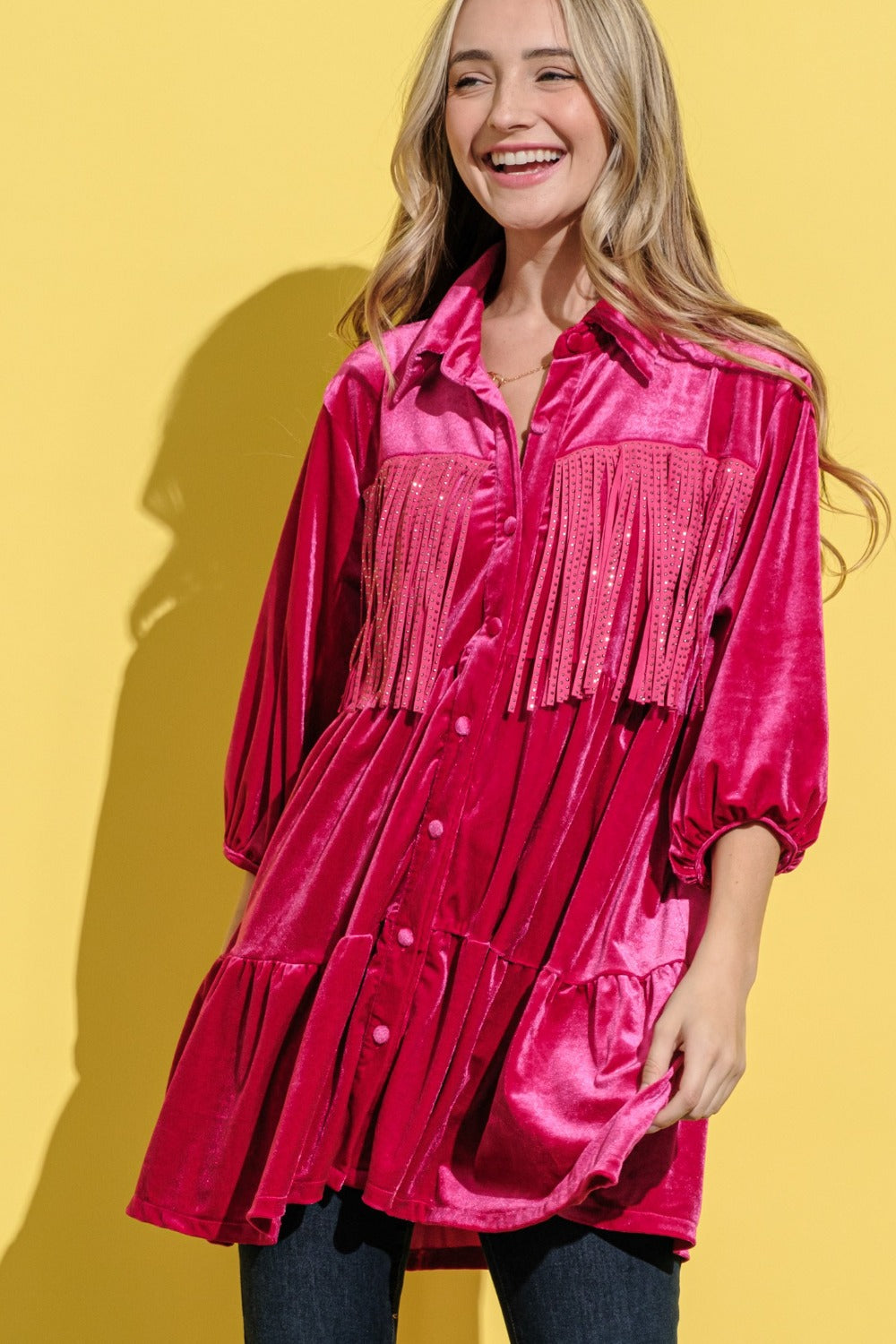 A woman stands against a bright yellow background, smiling and wearing an elegant statement fringed, bright pink And The Why Fringe Detailed Velvet Shirt Dress.