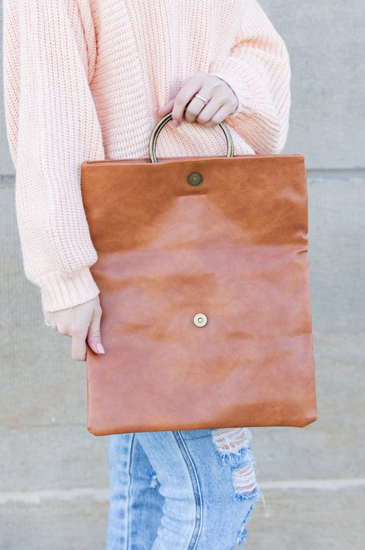 A person holds a tan Fold-over O-ring Clutch with a circular handle in front of their floral skirt and cream top.