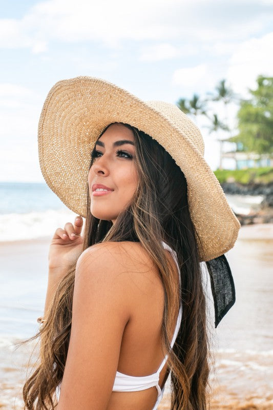 A woman wearing a white swimsuit holds onto her wide-brimmed, breathable Bow Accent Peasant Sun Hat at the beach.