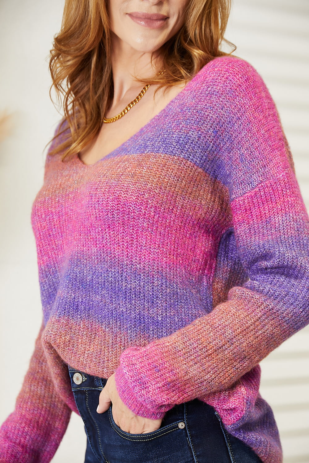 A woman with long, light brown hair stands indoors, smiling with her hands on her hips while wearing the Angel Wings Multicolored Rib-Knit V-Neck Knit Pullover paired with dark jeans. Beige curtains and a white piece of furniture are in the background.