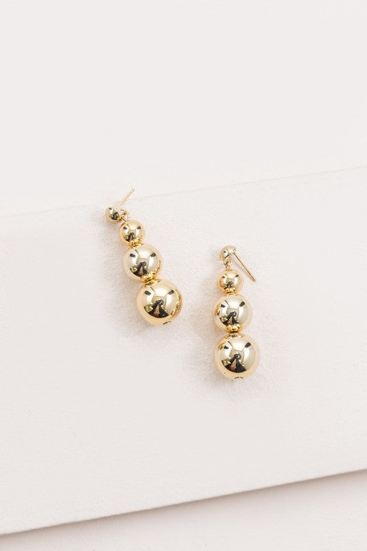 The Tiers in Heaven Golden Orb Earrings, featuring three connected gilded orbs, rest elegantly on a white surface.