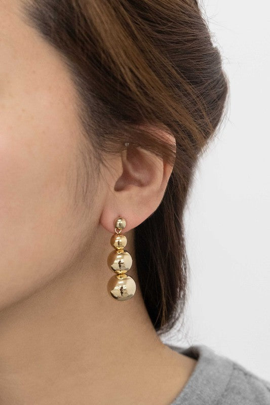 The Tiers in Heaven Golden Orb Earrings, featuring three connected gilded orbs, rest elegantly on a white surface.