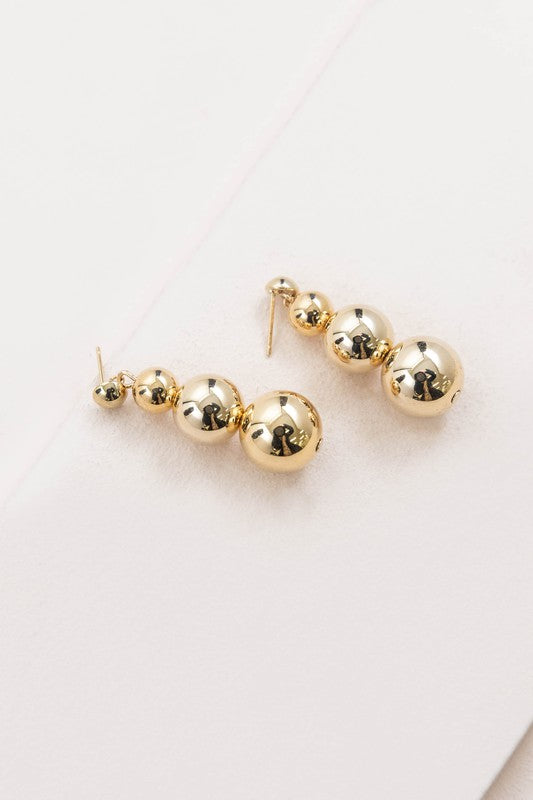 The Tiers in Heaven Golden Orb Earrings, featuring three connected gilded orbs, rest elegantly on a white surface.