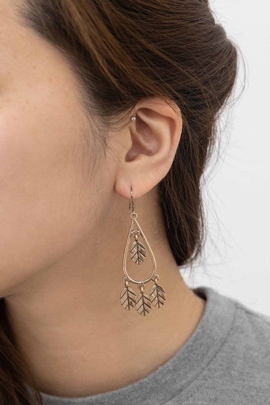 The Wind Swept Leaf Earrings showcase silver-toned teardrops embellished with numerous leaf-shaped pendants, making a striking impression against a light background.