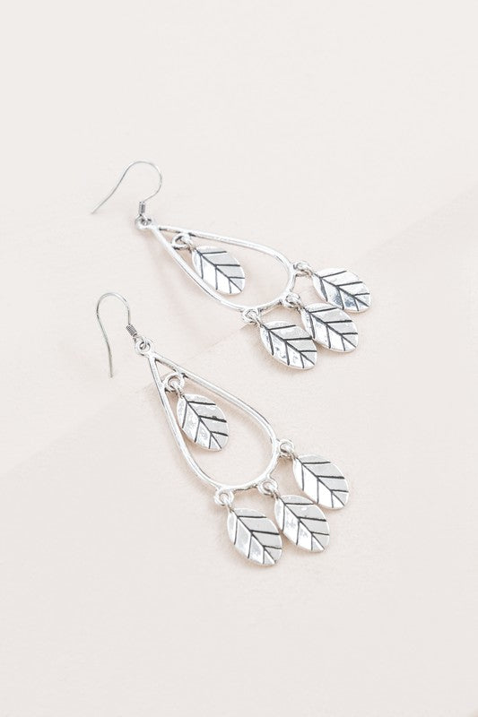 The Wind Swept Leaf Earrings showcase silver-toned teardrops embellished with numerous leaf-shaped pendants, making a striking impression against a light background.