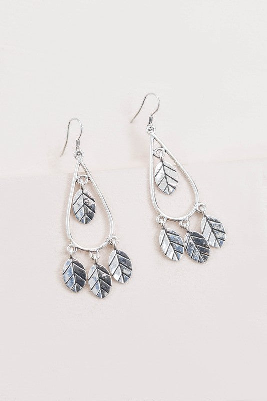 The Wind Swept Leaf Earrings showcase silver-toned teardrops embellished with numerous leaf-shaped pendants, making a striking impression against a light background.