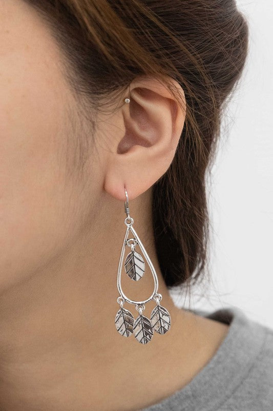 The Wind Swept Leaf Earrings showcase silver-toned teardrops embellished with numerous leaf-shaped pendants, making a striking impression against a light background.