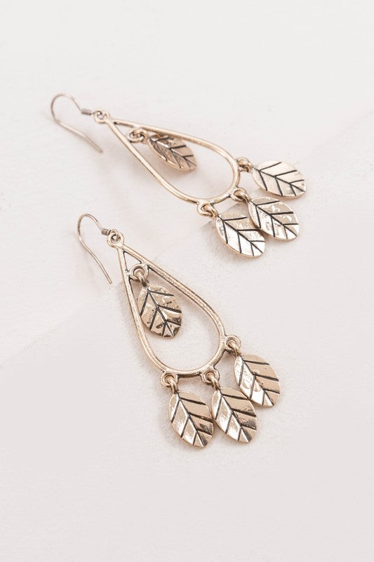 The Wind Swept Leaf Earrings showcase silver-toned teardrops embellished with numerous leaf-shaped pendants, making a striking impression against a light background.