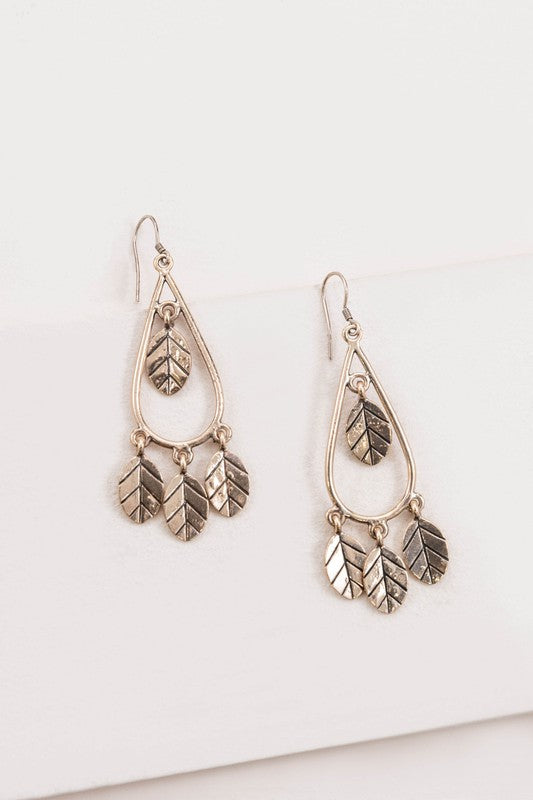 The Wind Swept Leaf Earrings showcase silver-toned teardrops embellished with numerous leaf-shaped pendants, making a striking impression against a light background.