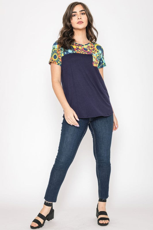 A woman smiles with her hands clasped, dressed in the Medallion Front Pocket Tunic, featuring a dark blue base with colorful, patterned sleeves.