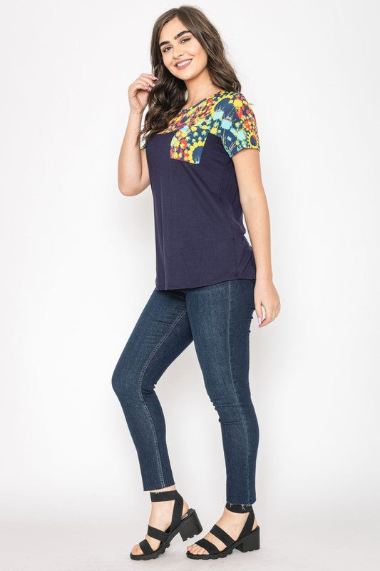 A woman smiles with her hands clasped, dressed in the Medallion Front Pocket Tunic, featuring a dark blue base with colorful, patterned sleeves.