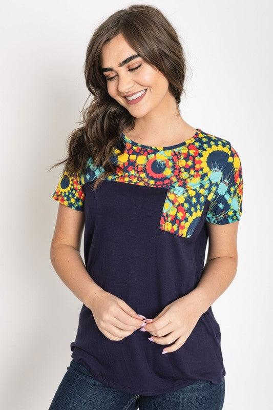 A woman smiles with her hands clasped, dressed in the Medallion Front Pocket Tunic, featuring a dark blue base with colorful, patterned sleeves.