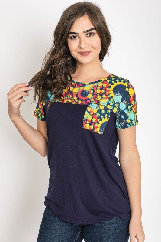 A woman smiles with her hands clasped, dressed in the Medallion Front Pocket Tunic, featuring a dark blue base with colorful, patterned sleeves.