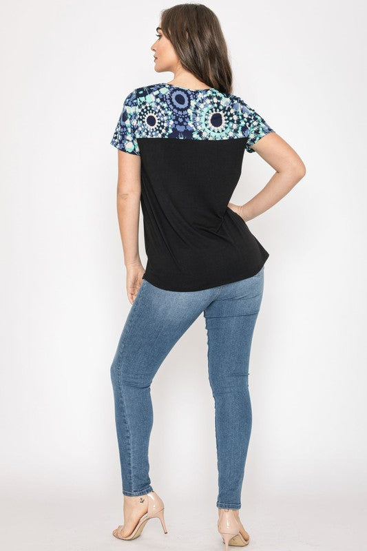 A woman wears the Plus Medallion Front Pocket Tunic, highlighted by a black lower section and a multicolored medallion-patterned upper part. She stands against a plain background, emphasizing the tunic's unique style.