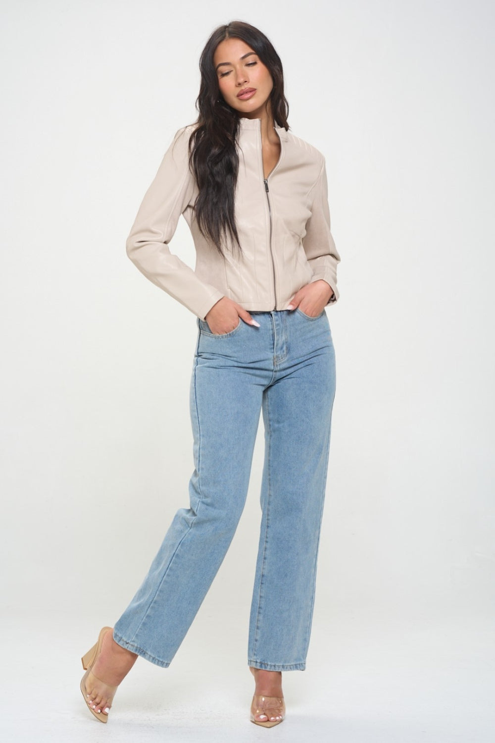 A woman with long dark hair, wearing the cruelty-free Coalition LA Zip Up Vegan Moto Jacket in light beige, pairs it with blue jeans as she stands with her arms crossed.