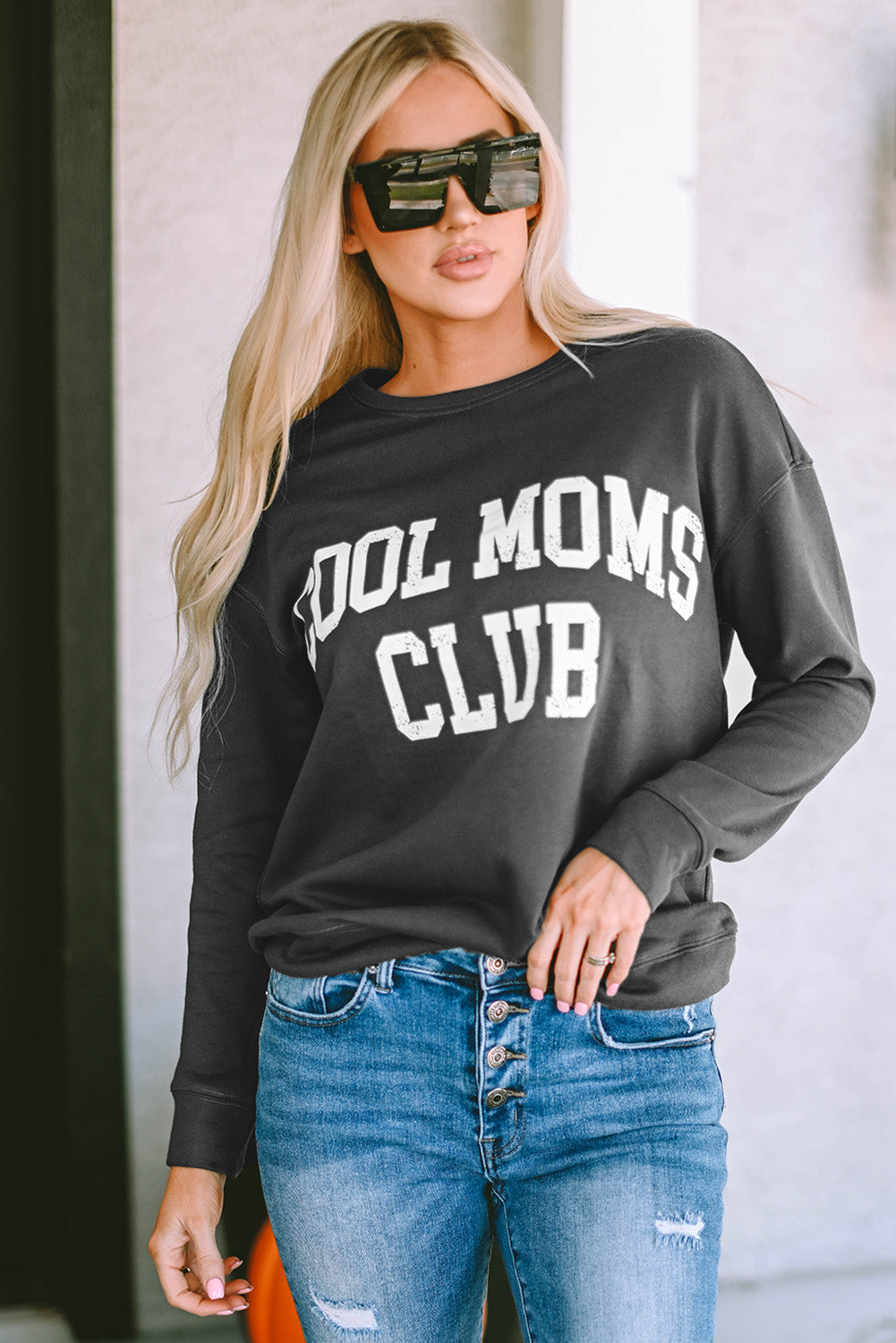 A person with long blond hair wearing a gray COOL MOMS CLUB drop shoulder sweatshirt and relaxed-fit blue jeans is seen from the back, standing near a pumpkin and a window.