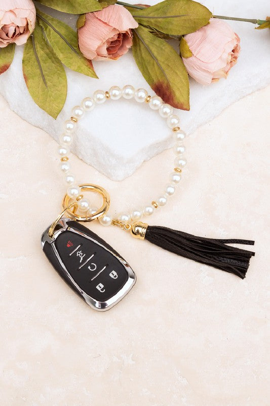 A person holding a Classic Pearl Key Ring Bracelet accessorized with a gold key ring bracelet, vegan leather tassel, and a car key.