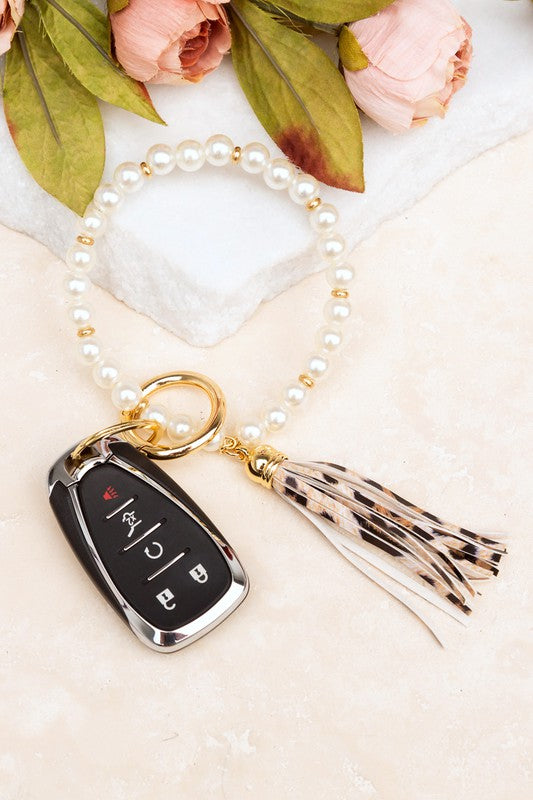 A person holding a Classic Pearl Key Ring Bracelet accessorized with a gold key ring bracelet, vegan leather tassel, and a car key.