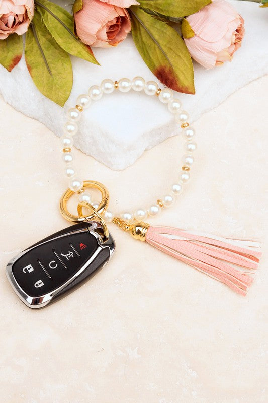 A person holding a Classic Pearl Key Ring Bracelet accessorized with a gold key ring bracelet, vegan leather tassel, and a car key.