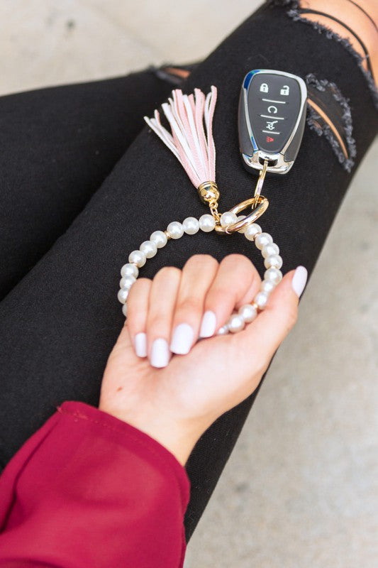 A person holding a Classic Pearl Key Ring Bracelet accessorized with a gold key ring bracelet, vegan leather tassel, and a car key.