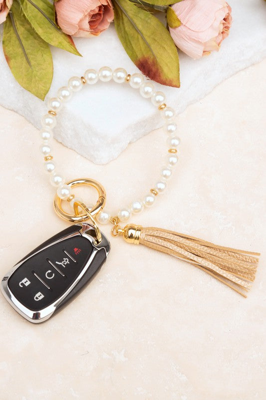 A person holding a Classic Pearl Key Ring Bracelet accessorized with a gold key ring bracelet, vegan leather tassel, and a car key.
