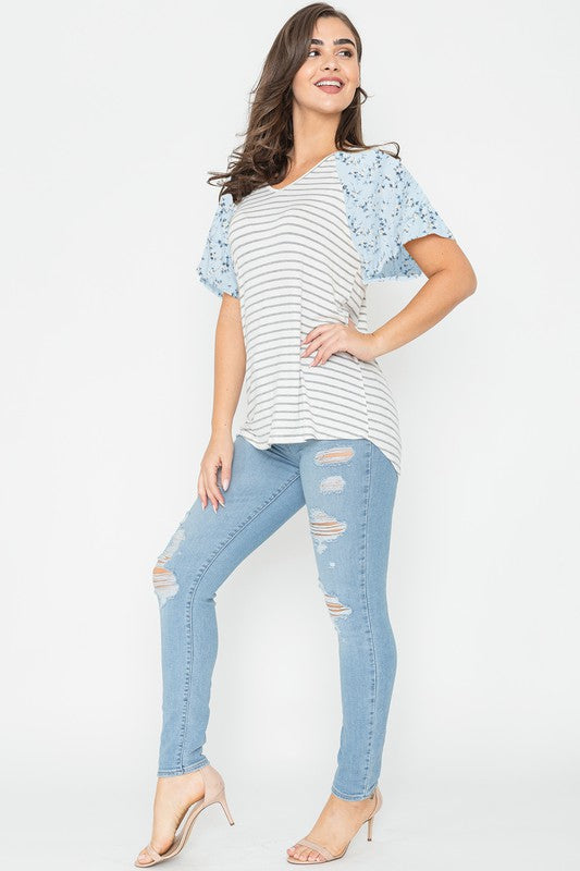 A woman is standing and smiling in a V Neck Stripe Floral Sleeve Tunic, paired perfectly with distressed jeans.