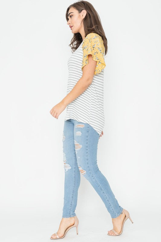 A woman is standing and smiling in a V Neck Stripe Floral Sleeve Tunic, paired perfectly with distressed jeans.