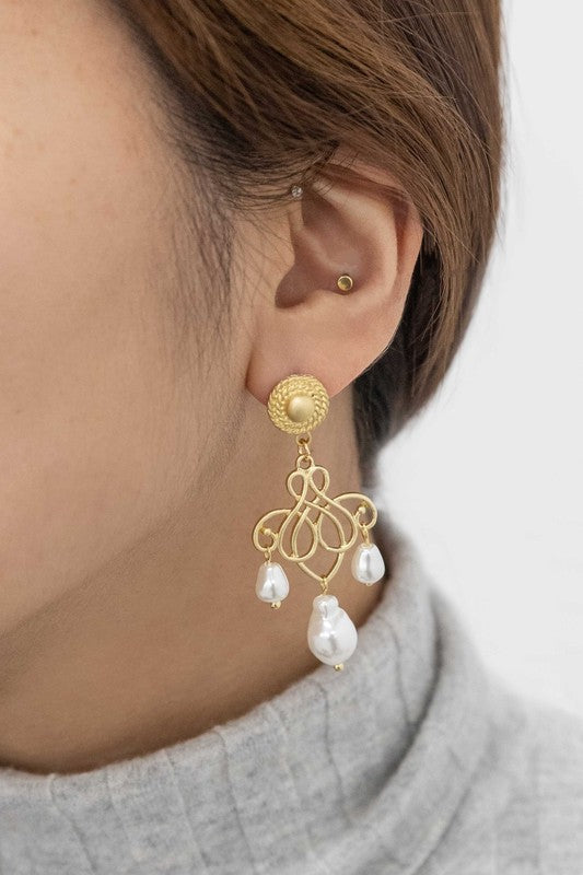 The Candelabrum Drop Earrings, featuring an intricate heart-shaped design and pearl dangles, evoke a formal style as they are beautifully displayed on a soft beige surface.