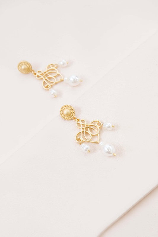 The Candelabrum Drop Earrings, featuring an intricate heart-shaped design and pearl dangles, evoke a formal style as they are beautifully displayed on a soft beige surface.