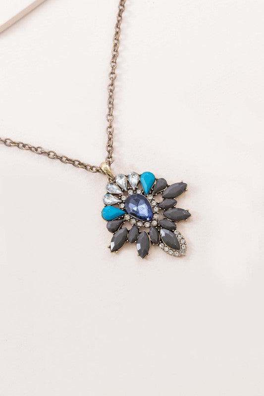 A Sapphira Pendant Necklace featuring a mix of blue and turquoise gemstones and rhinestones on a chain with a lobster clasp, against a neutral background.