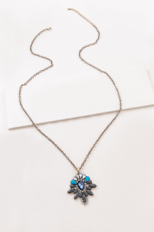 A Sapphira Pendant Necklace featuring a mix of blue and turquoise gemstones and rhinestones on a chain with a lobster clasp, against a neutral background.