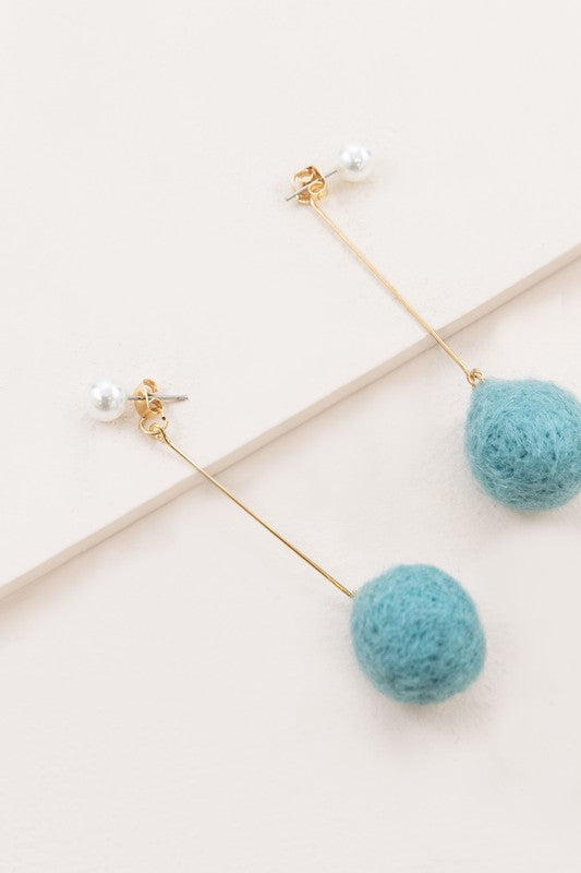 The Tess Pom Earrings are a pair of casual pearl earrings, showcasing long gold stems with small pearls at the top and sea blue pom spheres dangling at the bottom, elegantly displayed on a light surface.