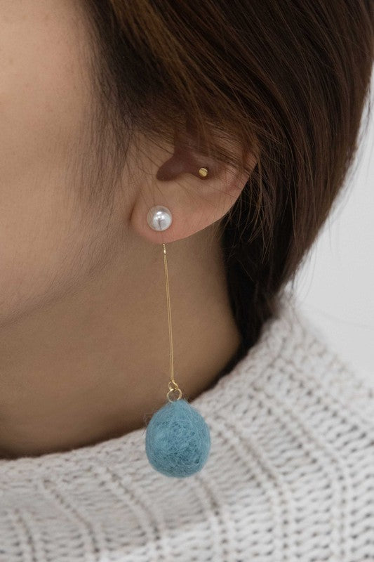 The Tess Pom Earrings are a pair of casual pearl earrings, showcasing long gold stems with small pearls at the top and sea blue pom spheres dangling at the bottom, elegantly displayed on a light surface.