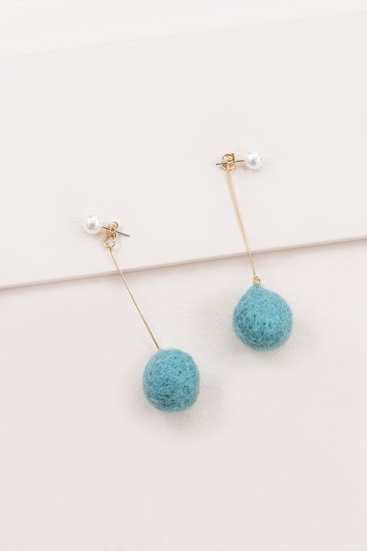 The Tess Pom Earrings are a pair of casual pearl earrings, showcasing long gold stems with small pearls at the top and sea blue pom spheres dangling at the bottom, elegantly displayed on a light surface.