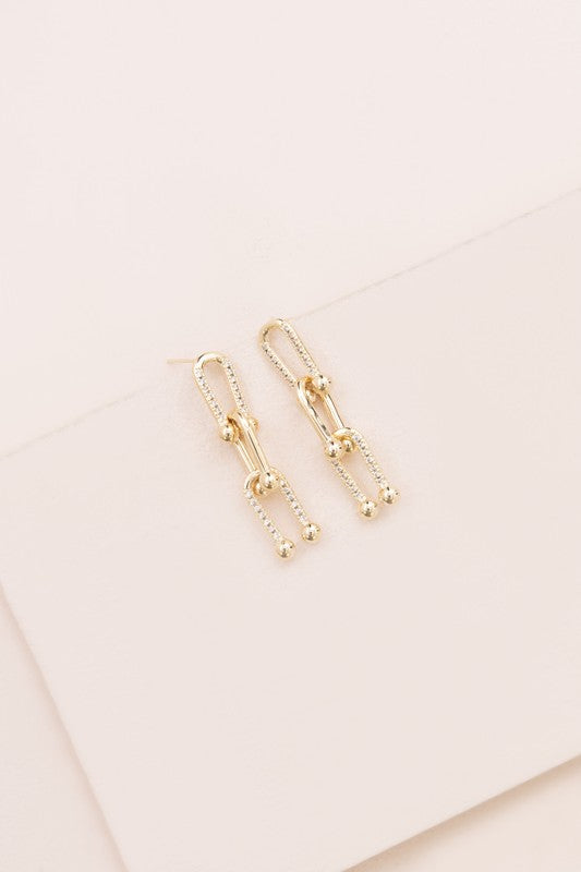 The U Link Drop Earrings, adorned with a geometric design and delicate pave stones, are crafted with 14k gold plating and displayed on a light-colored surface.