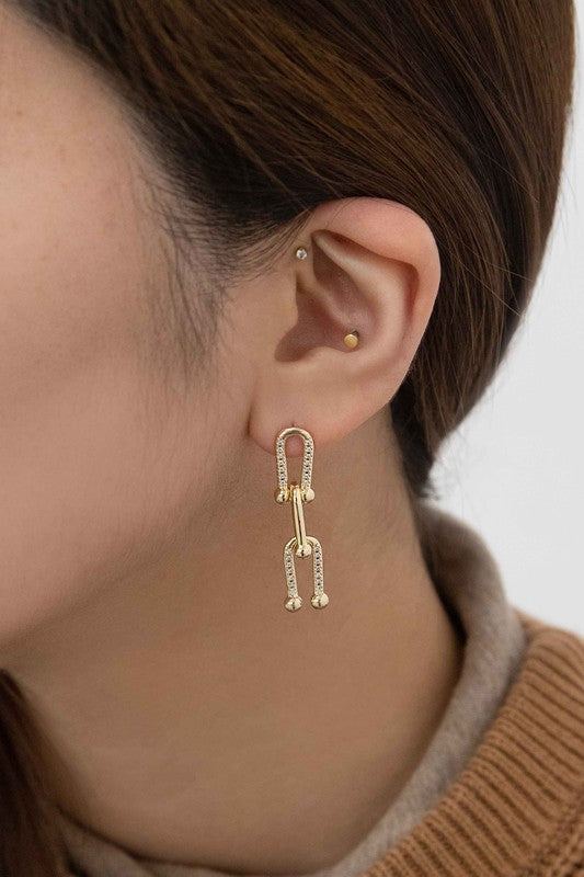 The U Link Drop Earrings, adorned with a geometric design and delicate pave stones, are crafted with 14k gold plating and displayed on a light-colored surface.