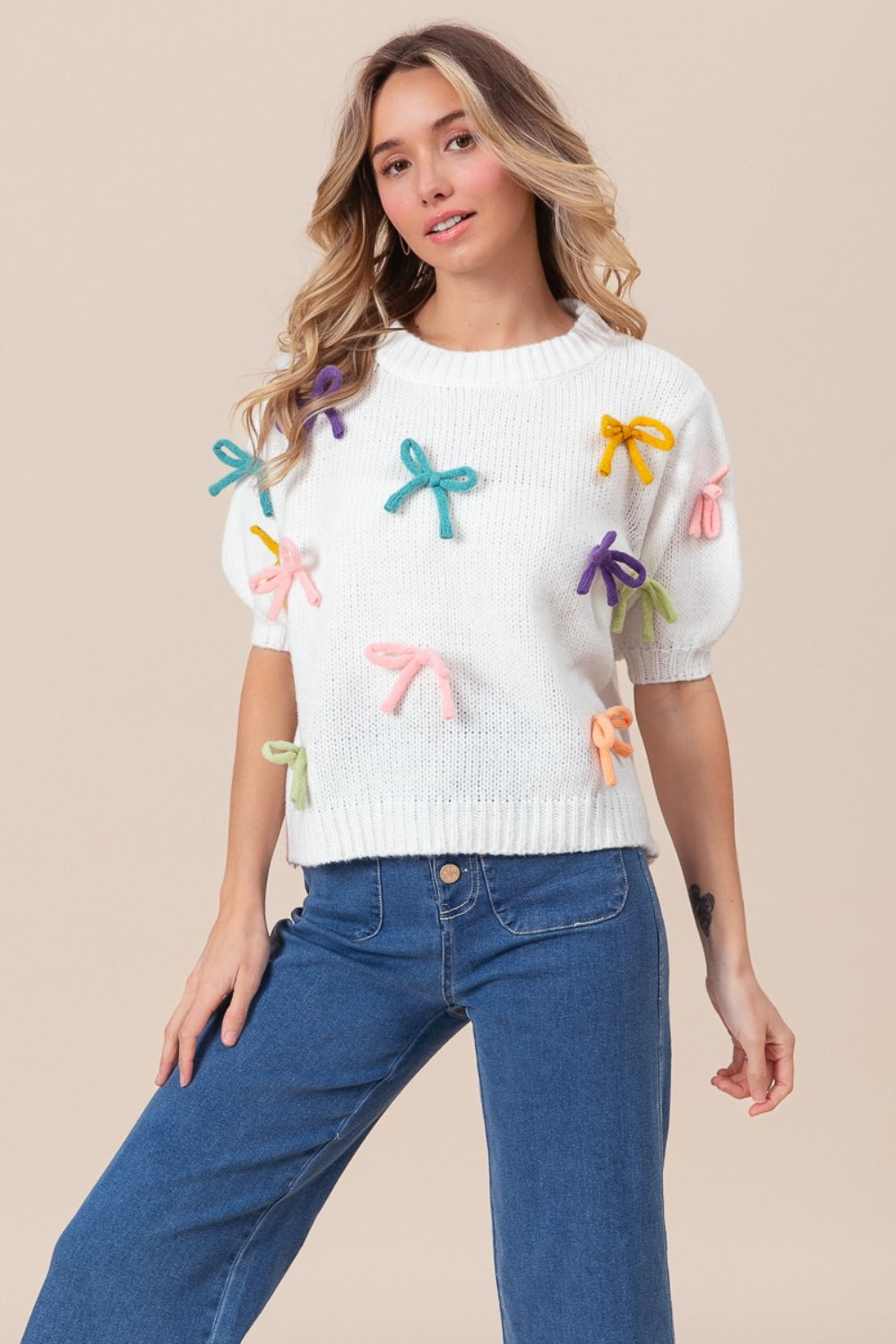 A woman stands against a beige background, wearing the BiBi Bow Detail Puff Sleeve Sweater in cozy white fabric with colorful bow decorations, paired with blue jeans.