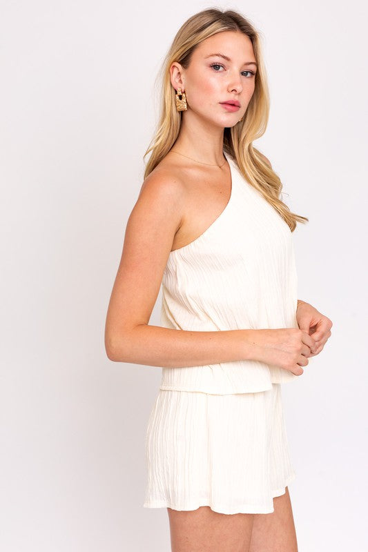 A person exudes contemporary elegance, dressed in a sleeveless one-shoulder layered top romper in white while effortlessly carrying a brown handbag.