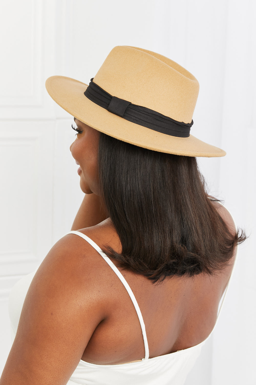 A woman in a white dress with adjustable straps, paired with the dark tan Fame You Got It Fedora Hat featuring a black band, looks over her shoulder and smiles.