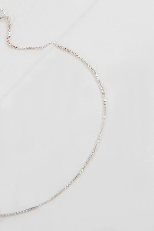 The Ana Plain Chain Anklet, ideal for casual jewelry enthusiasts, showcases a dainty silver chain with a straightforward clasp and adjustable extension links, elegantly laid out on a light gray surface.