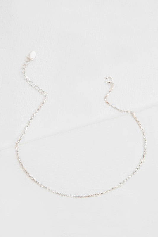 The Ana Plain Chain Anklet, ideal for casual jewelry enthusiasts, showcases a dainty silver chain with a straightforward clasp and adjustable extension links, elegantly laid out on a light gray surface.