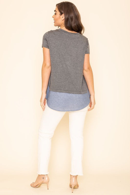 A woman wearing a Short Sleeve Layered Top stands against a beige background, highlighting the simplicity and comfort of this grey T-shirt with blue trim at the bottom, proudly made in the United States.