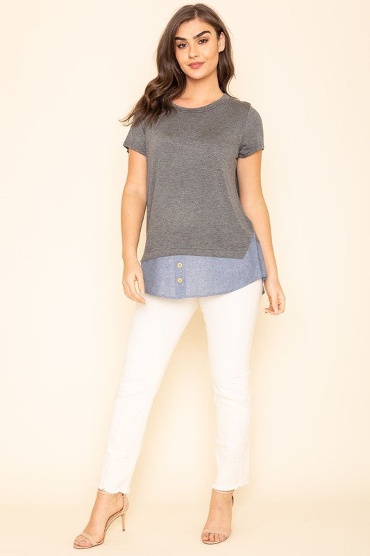 A woman wearing a Short Sleeve Layered Top stands against a beige background, highlighting the simplicity and comfort of this grey T-shirt with blue trim at the bottom, proudly made in the United States.